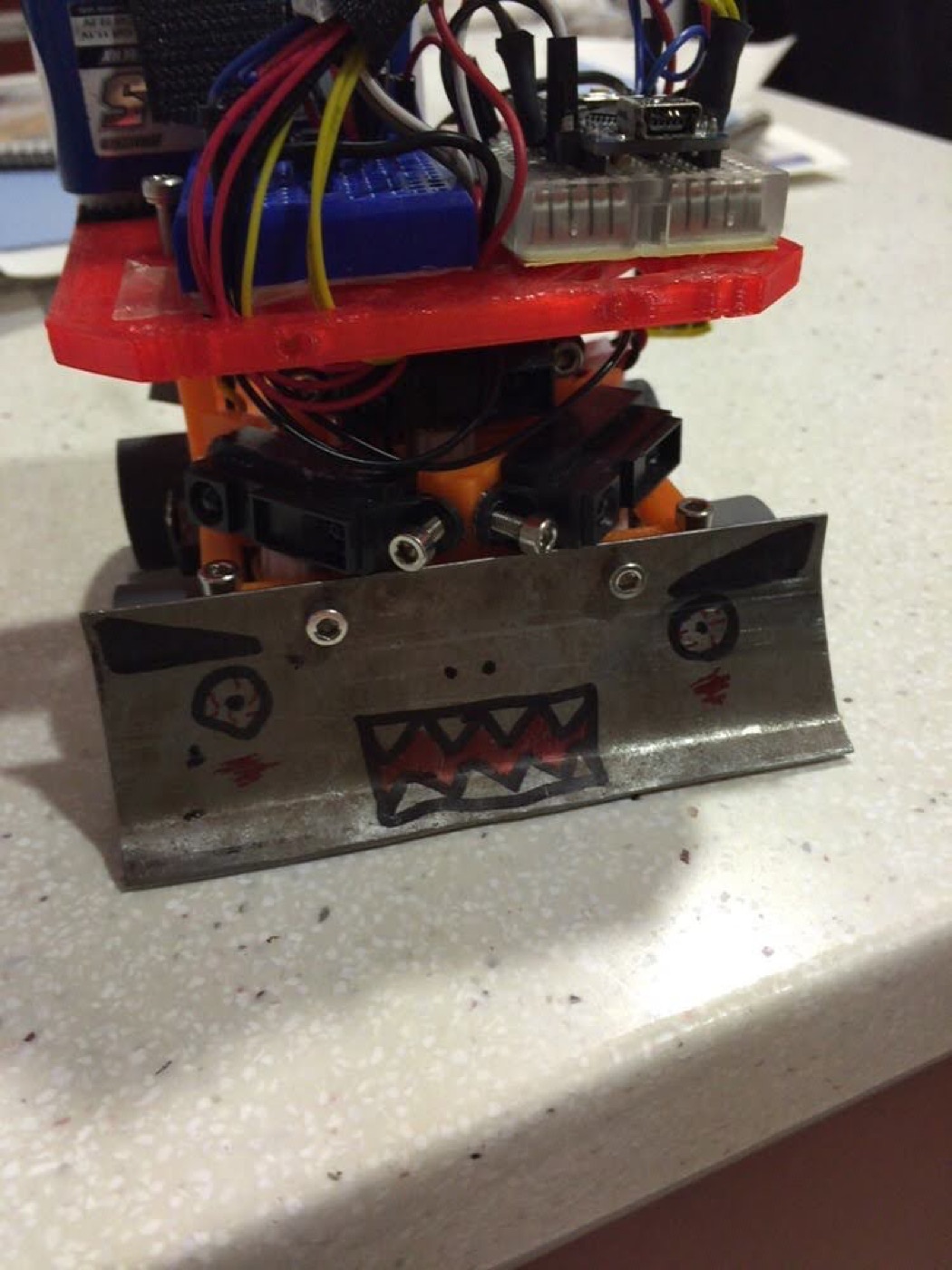 Minisumo visto de frente: una pala de acero con una cara dibujada, servos y una placa con cables encima