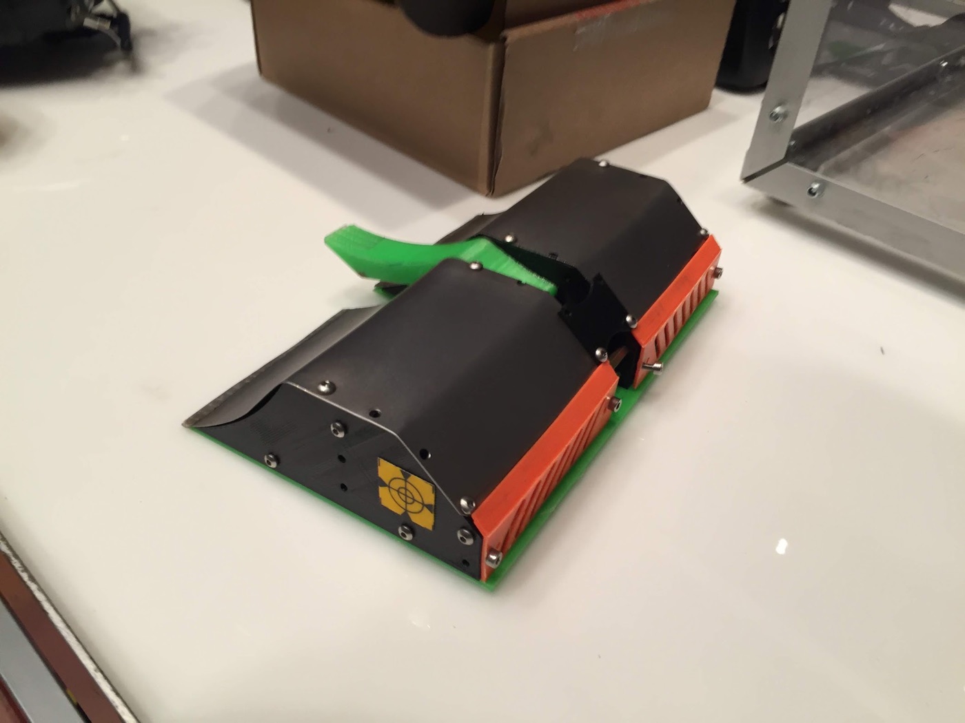 Robot volteador bajo con carcasa negra, laterales naranjas y la pala verde asomando por detrás