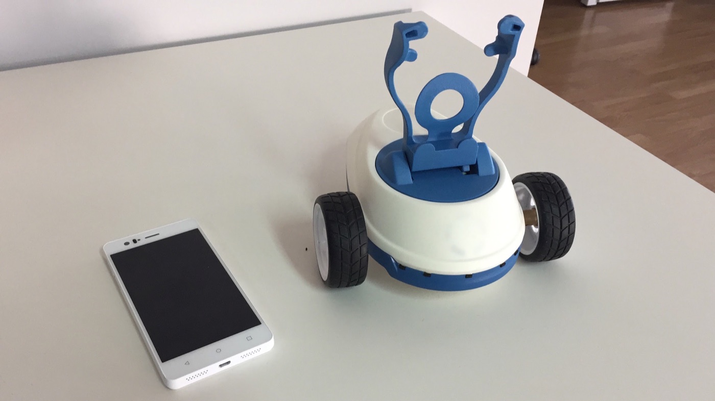 Base de Robobo blanca y azul con ruedas y un soporte vacío para el teléfono, junto a un smartphone sobre una mesa