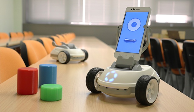 Robot Robobo sobre una mesa de aula: una base con ruedas y un smartphone que muestra una cara animada, junto a cilindros de colores y otro Robobo al fondo
